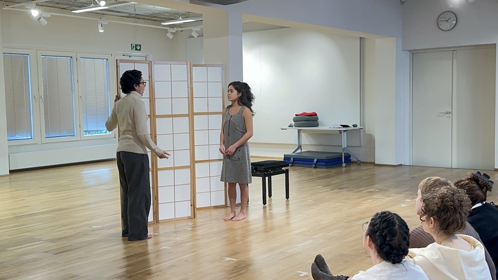 Two actors stand facing each other in mid-performance of 'Faith Hope Charity'. Both actors are standing  in front of a folding room divider in a bright acting studio. A small group of students sit watching them to the bottom right of the photo.
